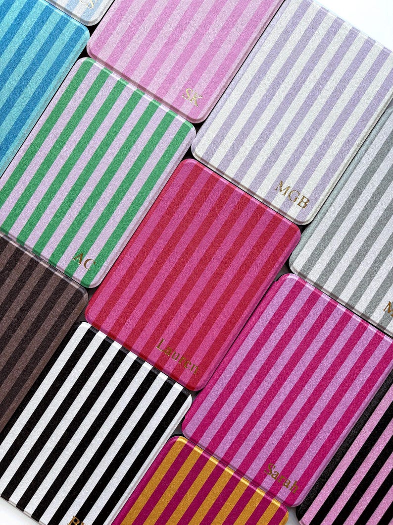 May include: A selection of rectangular cases with vertical stripes in various colours, including pink, green, black, and gold. Some cases have initials or names in gold lettering. The cases are arranged diagonally.