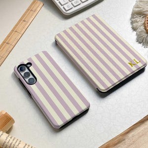 May include: Two phone cases and a phone case cover with a vertical striped pattern in lavender and cream. The cover has the initials "KL" in gold. A calculator, ruler, and a book are also visible.
