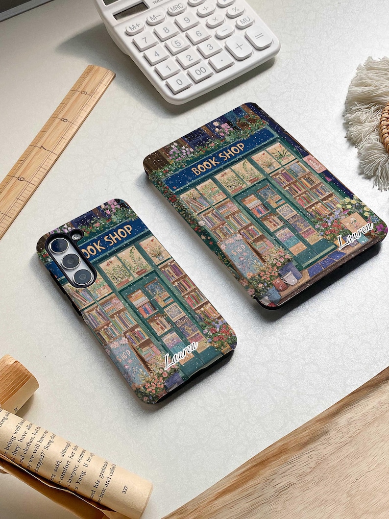May include: Two phone cases with a book shop illustration. The cases are black with a green, blue, and white illustration of a book shop with the text "BOOK SHOP" above the door. The text "Lauren" is written in white on the bottom of each case.