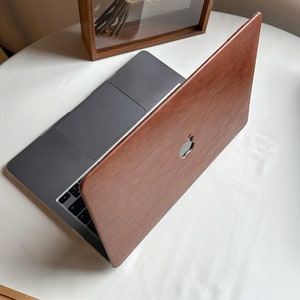 May include: A brown leather laptop case with a silver Apple logo. The case is closed and the laptop is visible inside.