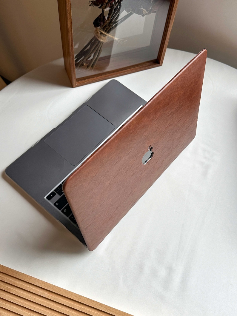 May include: A brown leather laptop case with a silver Apple logo. The case is closed and the laptop is visible inside.