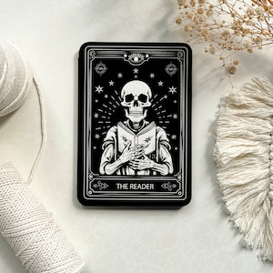 May include: Black and white tarot card with a skeleton holding a book. The card is titled "The Reader".