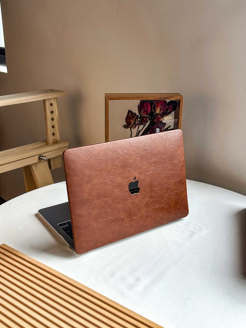 May include: A brown leather laptop case with the Apple logo on the front. The case is on a white table with a wooden slatted surface in the foreground.