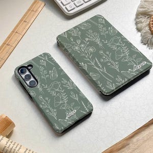 Puede incluir: Dos fundas para teléfono con fondo verde y diseños florales blancos. Las fundas tienen el nombre "Lauren" impreso.
