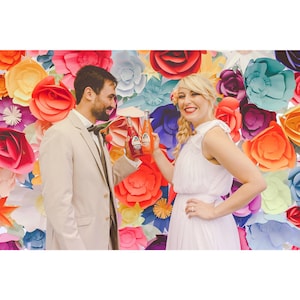 May include: A couple smiles, holding up bottles of soda in front of a colorful wall of paper flowers. The woman wears a white dress, and the man wears a tan suit. The background features large, vibrant paper flowers in various colors.
