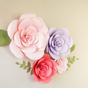 May include: A collection of large, handcrafted paper flowers in shades of pink, red, and purple, with green leaves and stems. The flowers are arranged on a light-colored background, creating a decorative floral display.