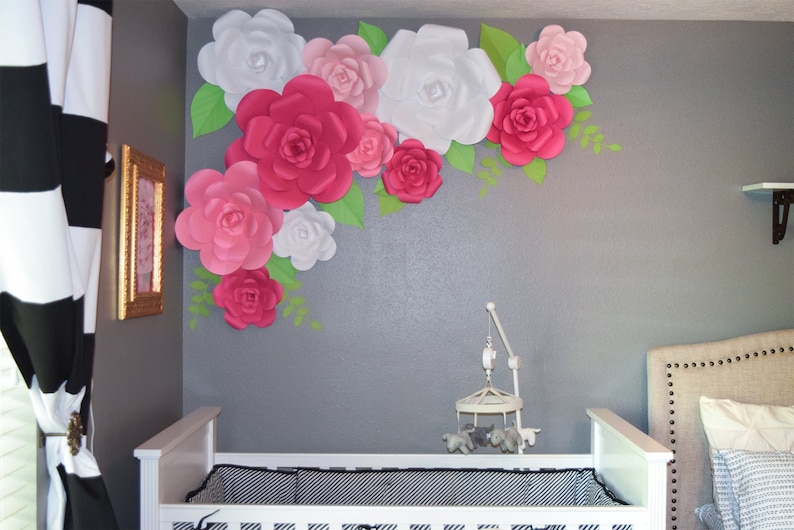 May include: A nursery with a white crib and a wall decorated with large paper flowers. The flowers are in shades of pink, white, and red, with green leaves. A mobile hangs above the crib.