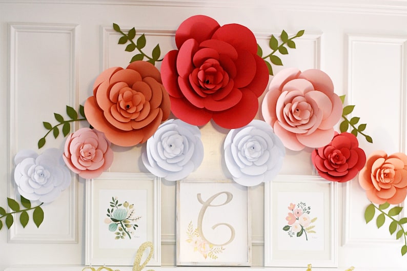 May include: A decorative arrangement featuring large paper flowers in shades of red, peach, pink, and white, accented with green leaf vines. Three framed floral prints are displayed below the flowers, creating a cohesive and artistic display.