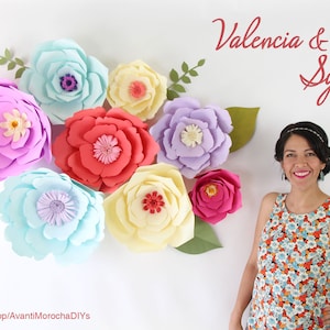 May include: A woman stands in front of a white wall decorated with large paper flowers in various colors, including pink, red, yellow, and blue. The flowers are arranged in a semi-circle around her. The text "Valencia & Sydney DIY" is written in red at the top of the image. The text "www.etsy.com/shop/AvantiMorochaDIYs" is written at the bottom of the image.