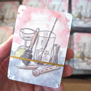 May include: Watercolor illustration of cocktail-making tools on a card. The image features a cocktail shaker, mixing glass, bar spoon, and other bar tools. The background has pink and blue watercolor washes, and the cards are held together with a gold rubber band.