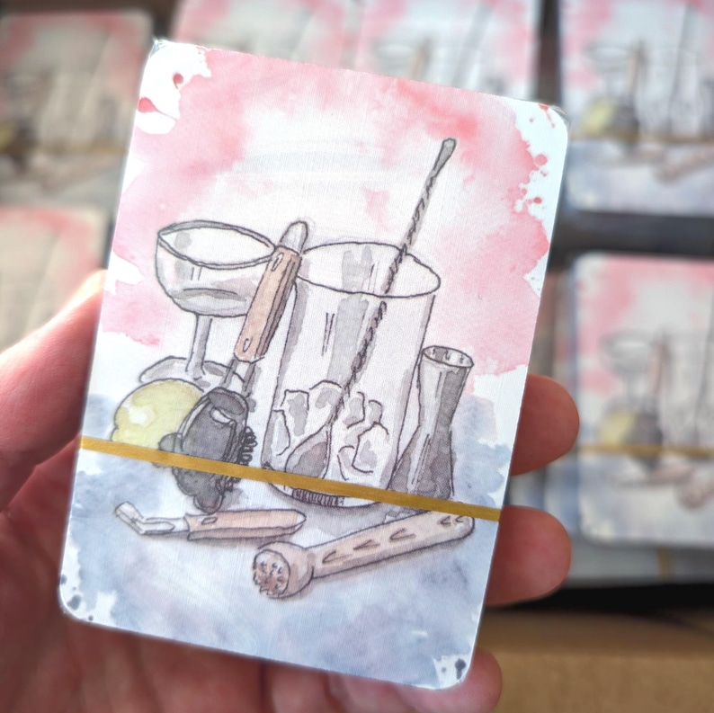May include: Watercolor illustration of cocktail-making tools on a card. The image features a cocktail shaker, mixing glass, bar spoon, and other bar tools. The background has pink and blue watercolor washes, and the cards are held together with a gold rubber band.