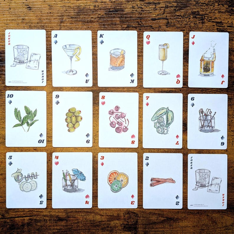May include: A deck of playing cards with watercolor illustrations of cocktails and garnishes. Each card features a different drink, such as a martini, old fashioned, and a highball. The cards are arranged on a wooden surface.
