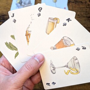 May include: A hand holding a deck of playing cards with cocktail illustrations. The cards feature the Ace, King, Queen, Jack, and 10 of Spades. Each card showcases a different cocktail with garnishes, in a watercolor style.