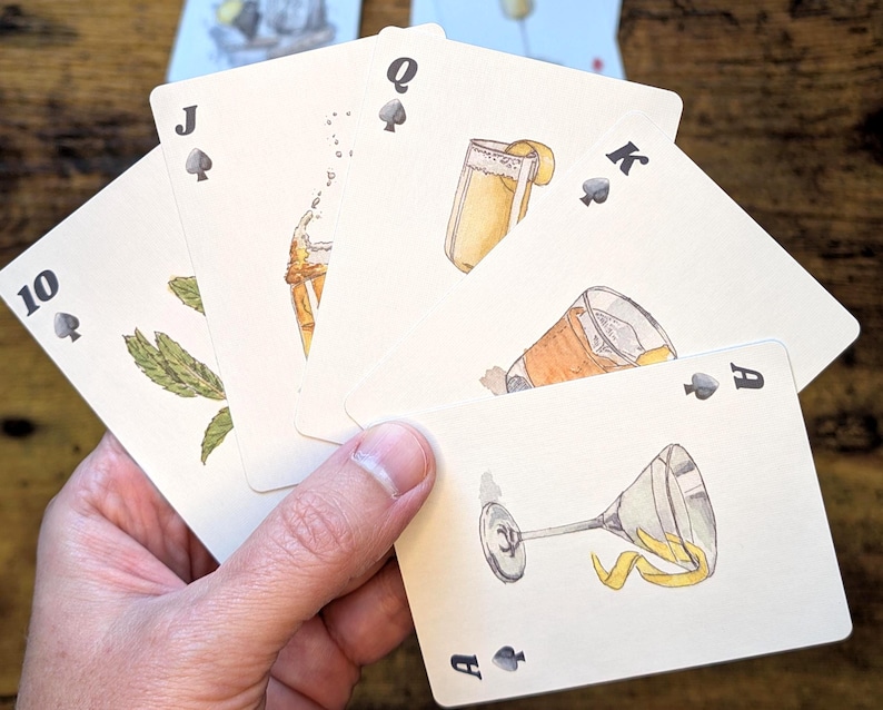 May include: A hand holding a deck of playing cards with cocktail illustrations. The cards feature the Ace, King, Queen, Jack, and 10 of Spades. Each card showcases a different cocktail with garnishes, in a watercolor style.