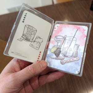 May include: A clear plastic playing card case holding two cards with cocktail-themed illustrations. One card features a drink with ice and a receipt, the other shows bar tools and a drink. The word "JOKER" is printed on the cards.