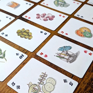 May include: A collection of playing cards with watercolor illustrations of cocktail ingredients. The cards feature images of olives, cherries, and citrus fruits. The cards are arranged on a wooden surface.