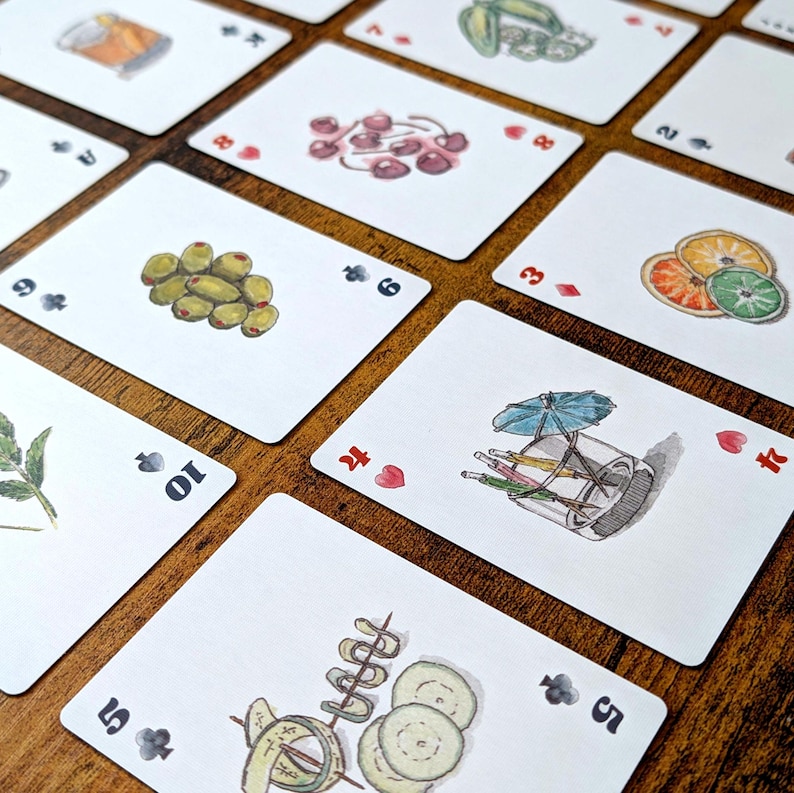 May include: A collection of playing cards with watercolor illustrations of cocktail ingredients. The cards feature images of olives, cherries, and citrus fruits. The cards are arranged on a wooden surface.