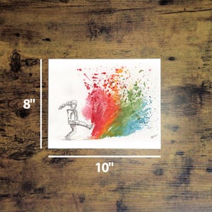 May include: An 8" x 10" watercolor print on a wooden surface. The artwork depicts a person kicking a colorful rainbow splash. The rainbow transitions from red to orange, yellow, green, and blue. The person is drawn in black and white.