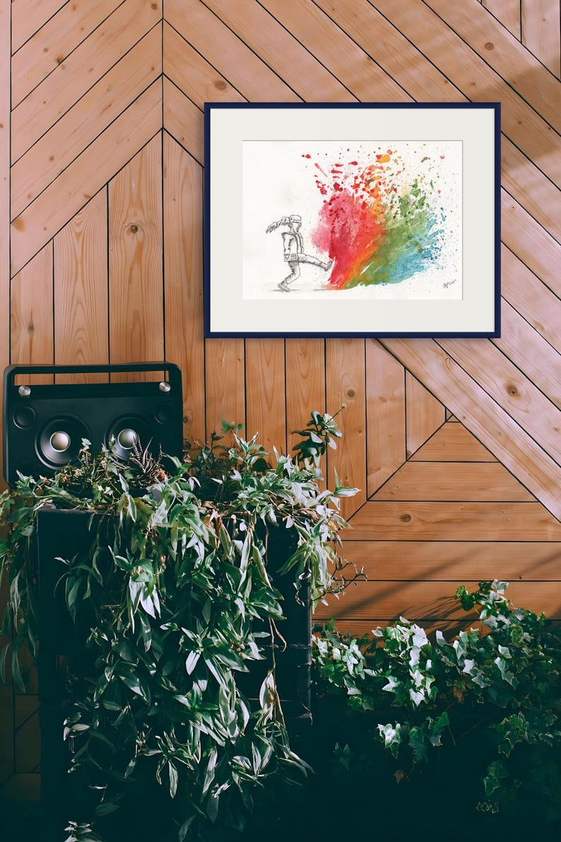 May include: A framed art print featuring a black and white sketch of a person kicking a colorful splash of paint. The artwork is set against a wooden wall, with a black speaker and greenery in the foreground.