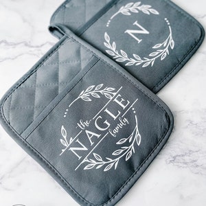 May include: Two gray quilted pot holders with white text and designs. One features the letter "N" inside a wreath, the other reads "the NAGLE family" within a similar wreath design. The pot holders are on a white marble surface.