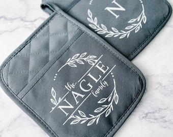 May include: Two gray quilted pot holders with white text and designs. One features the letter "N" inside a wreath, the other reads "the NAGLE family" within a similar wreath design. The pot holders are on a white marble surface.