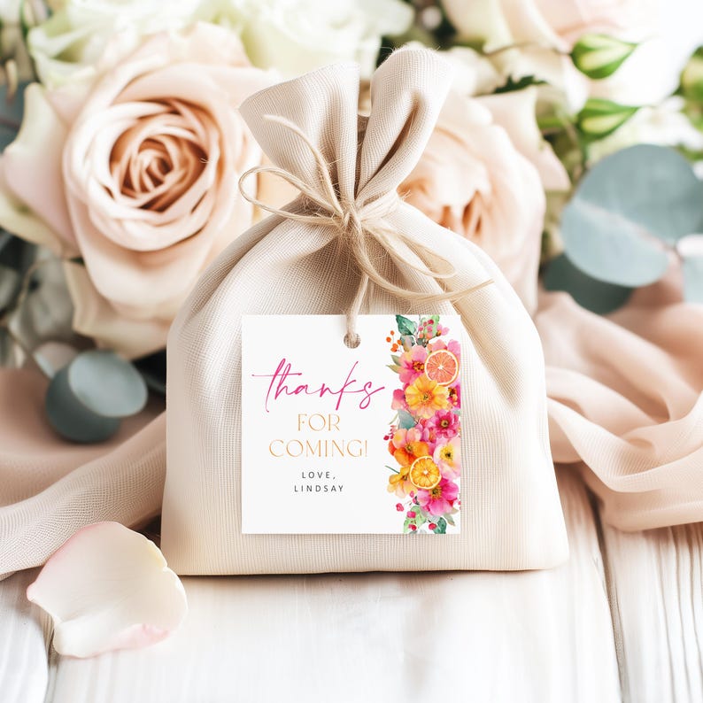 May include: A beige fabric gift bag tied with twine, with a white tag that reads "thanks for coming!" in pink and gold lettering. The tag is decorated with a floral design in shades of pink, orange, and yellow. The background features soft pink roses.