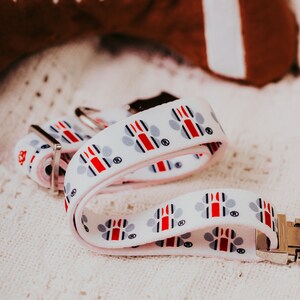 May include: White dog collar with red, blue and grey paw prints. The collar has a silver buckle and is lying on a white crocheted surface.