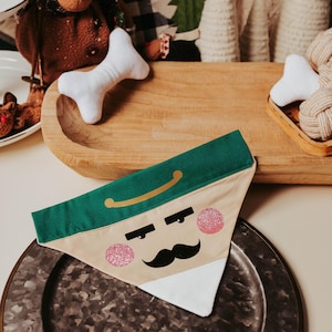 May include: A festive dog bandana with a nutcracker face design. The bandana features a green top, a tan face, a black mustache, and pink glitter cheeks. It is placed on a metal plate, with other holiday decorations in the background.