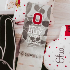 May include: A white and grey fabric tag with a red "O" and the text "Buckeyes Buy Buckeye. Officially Licensed Ohio State Product".