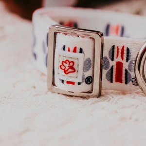 May include: White dog collar with a silver buckle and paw print design. The collar has red, blue and grey accents.