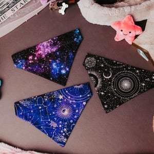 May include: Three triangular pet bandanas featuring celestial themes. One displays a colorful galaxy print, another shows a black and white sun and moon pattern, and the third features a blue background with white zodiac constellations and symbols.