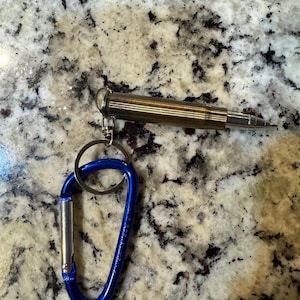 May include: A gold-colored metal keychain with a blue carabiner clip. The keychain is shaped like a bullet and has a small hole at the top for attaching to a keyring.