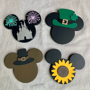 May include: Four wooden Mickey Mouse ears with different holiday themes. The first pair has a black background with a silver silhouette of Cinderella's castle and green and pink fireworks. The second pair has a black background with a green leprechaun hat with a gold buckle and a shamrock. The third pair has a brown background with a black pilgrim hat with a gold buckle. The fourth pair has a black background with a yellow sunflower.