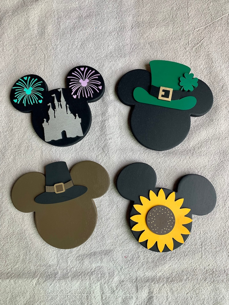 May include: Four wooden Mickey Mouse ears with different holiday themes. The first pair has a black background with a silver silhouette of Cinderella's castle and green and pink fireworks. The second pair has a black background with a green leprechaun hat with a gold buckle and a shamrock. The third pair has a brown background with a black pilgrim hat with a gold buckle. The fourth pair has a black background with a yellow sunflower.