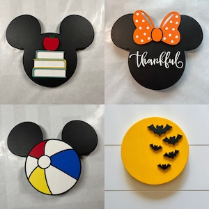 May include: Four different wooden ornaments featuring a Mickey Mouse silhouette. The first ornament is black with a red apple and stacked books. The second ornament is black with a white bow and the word "thankful". The third ornament is black with a beach ball. The fourth ornament is yellow with black bats.