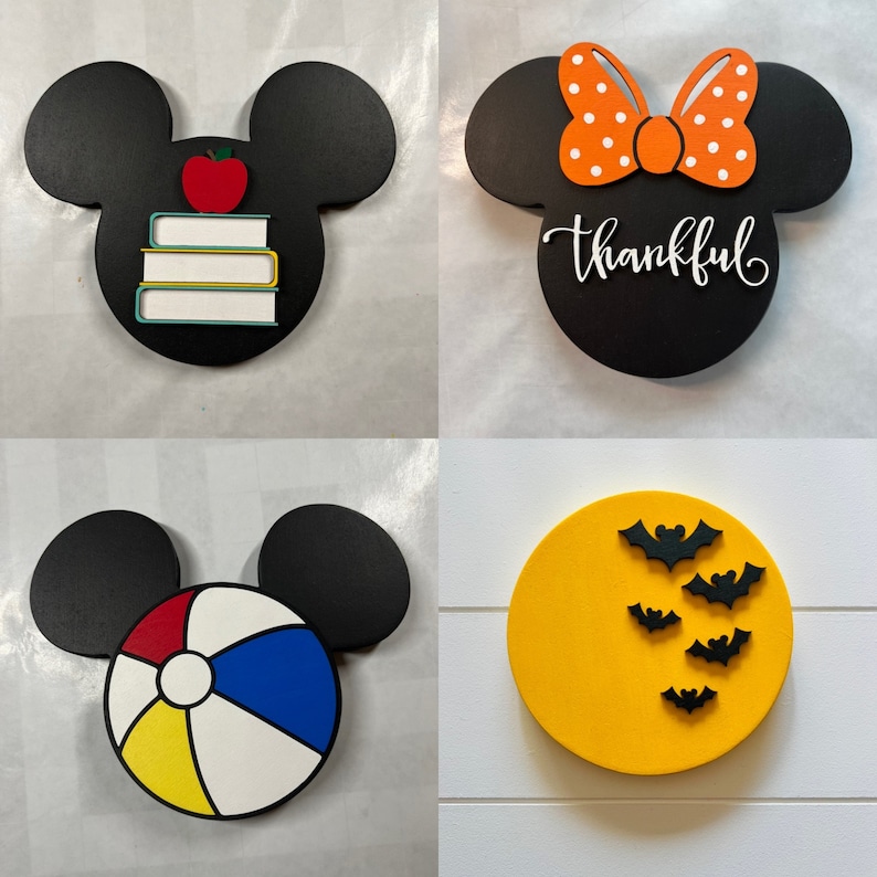 May include: Four different wooden ornaments featuring a Mickey Mouse silhouette. The first ornament is black with a red apple and stacked books. The second ornament is black with a white bow and the word "thankful". The third ornament is black with a beach ball. The fourth ornament is yellow with black bats.