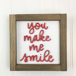 May include: A square wooden sign with a brown frame and a white background. The words "you make me smile" are in red, cursive lettering. The sign is a decorative item.