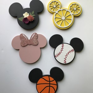 May include: Five wooden Mickey Mouse head shapes with different designs. One is black with a floral design, one is pink with a pink glitter bow, one is yellow with a lemon slice design, one is white with a baseball design, and one is black with a basketball design.