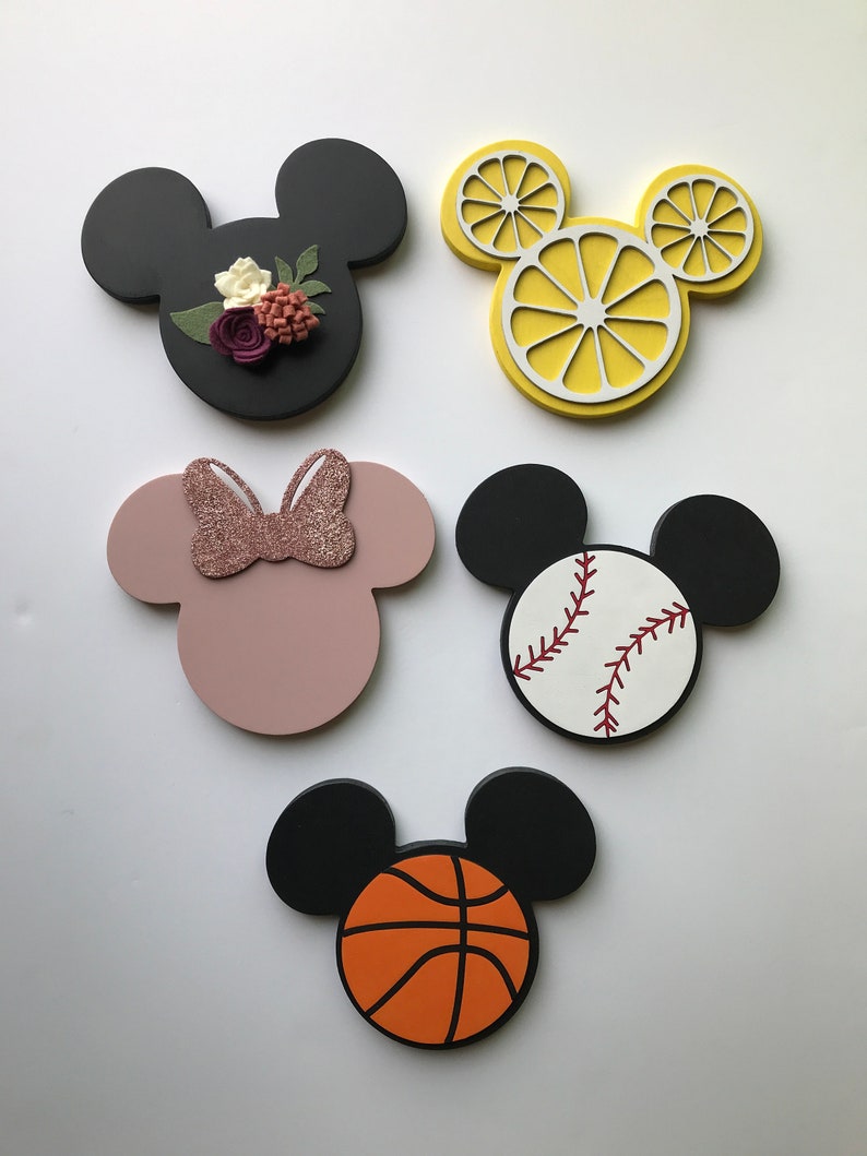 May include: Five wooden Mickey Mouse head shapes with different designs. One is black with a floral design, one is pink with a pink glitter bow, one is yellow with a lemon slice design, one is white with a baseball design, and one is black with a basketball design.