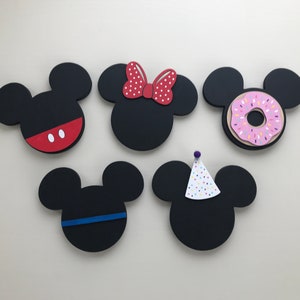 May include: Five black wooden Mickey Mouse head shapes with different decorations. One has red and white polka dot bow, one has a pink donut with sprinkles, one has a red and white polka dot bow, one has a blue stripe, and one has a white and colorful party hat.