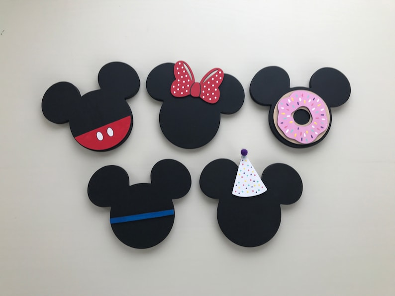May include: Five black wooden Mickey Mouse head shapes with different decorations. One has red and white polka dot bow, one has a pink donut with sprinkles, one has a red and white polka dot bow, one has a blue stripe, and one has a white and colorful party hat.