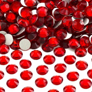 May include: Close-up of numerous red, faceted rhinestones with silver backing. The gemstones are scattered across a white surface, creating a sparkling, textured effect. Ideal for crafting, jewelry making, or embellishing various items.