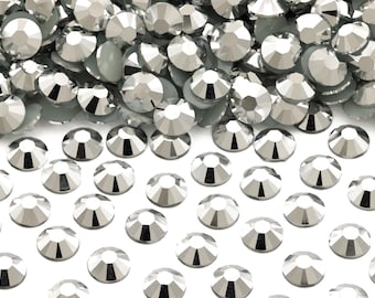 May include: A collection of silver rhinestones with a faceted design. The rhinestones are round with a flat back and a shiny, reflective surface. They are scattered across a white background.