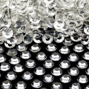 May include: A close-up image of clear, faceted rhinestones. The rhinestones are arranged in a random pattern on a black surface.