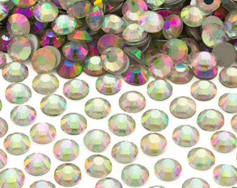May include: A collection of iridescent, round rhinestones with a flat back. The gems display a range of colors, including pink, purple, green, and yellow, and are scattered across a white surface. These decorative embellishments are suitable for various craft projects.