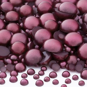 May include: A collection of burgundy-colored half-round beads, varying in size, scattered on a white surface. Some beads are whole, while others are cut in half, revealing a lighter interior. The beads have a glossy, pearl-like finish.