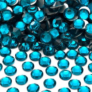 May include: A large pile of teal blue rhinestones, with a few scattered on a white surface.