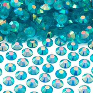 May include: A close-up shot showcases a collection of iridescent teal rhinestones. These small, faceted gems are scattered across a white surface, creating a sparkling and decorative effect. The rhinestones have a flat back and are ideal for embellishing crafts.