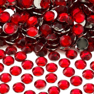 May include: A large pile of red, round, flat-backed rhinestones. The rhinestones are arranged in a random pattern and are scattered on a white surface.