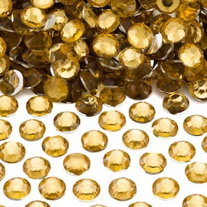 May include: A pile of gold-colored, faceted, round rhinestones. The rhinestones are scattered on a white surface.